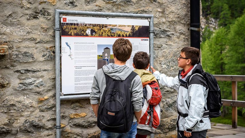 Tre ragazzi leggono un pannello informativo sulla centrale Palü durante una visita guidata.
