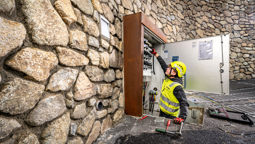 Netzelektriker arbeitet an einem offenen Schaltschrank in einer Steinmauer; Werkzeug und Kabel liegen bereit.