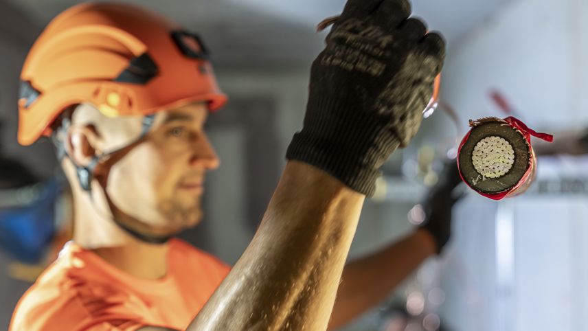 Monteur mit Helm hält den Querschnitt eines Stromkabels; im Hintergrund weitere Arbeiten an der Anlage.