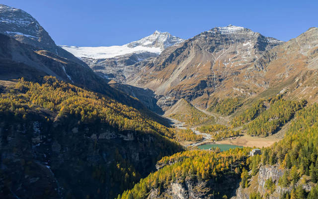 Blick über ein alpines Tal mit Gletscher, Lärchenwald und kleinem See in herbstlicher Berglandschaft.