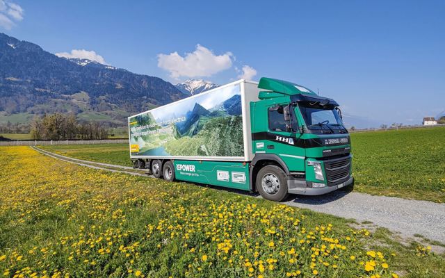 Elektro-LKW von Hunger fährt durch eine blühende Wiese vor alpiner Landschaft und blauem Himmel.
